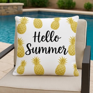 Può includere: Cuscino quadrato bianco con ananas gialli e la scritta "Hello Summer" in nero. Il cuscino è su una sedia beige vicino a una piscina.