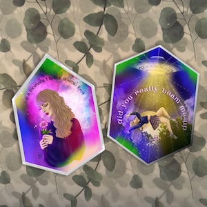 May include: Two hexagonal shaped, colorful, illustrated prints with white borders. The first print features a woman holding a pink flower with the text "for a moment I knew cosmic love". The second print features a woman floating in the air with a UFO above her and the text "did you really beam me up".