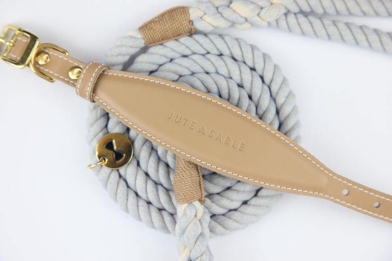 Pu&ograve; includere: Un set collare e guinzaglio per cani con un collare in pelle marrone chiaro e finiture color oro. Il collare &egrave; cucito con filo bianco e impresso con "JUTE & SABLE". Il guinzaglio e il collare sono su una corda grigia arrotolata.