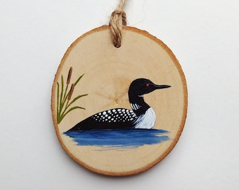 May include: A hand-painted wooden ornament featuring a loon swimming in blue water with green reeds. The ornament is attached to a twine string for hanging.