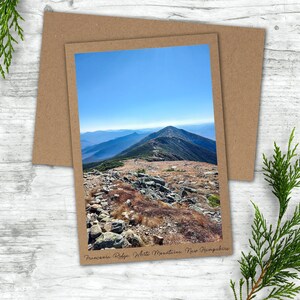 May include: A scenic view of Franconia Ridge in the White Mountains of New Hampshire. The photo shows a rocky mountain path with a blue sky and distant mountains in the background.