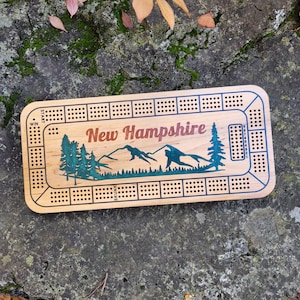 May include: Wooden cribbage board with the text "New Hampshire" in a rustic font. The board features a teal mountain and tree design. The board has multiple holes for pegs and is rectangular in shape.