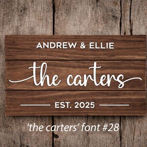 May include: A rectangular wooden sign with the names "Andrew & Ellie" above "the carters" in a cursive font. Below is "EST. 2025". The sign is brown with white lettering, set against a wooden background. The text "'the carters' font #28" is at the bottom.