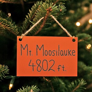 May include: An orange rectangular sign with the text "Mt. Moosilauke 4802 ft." is suspended from a Christmas tree. The sign is attached with twine and the background is blurred.