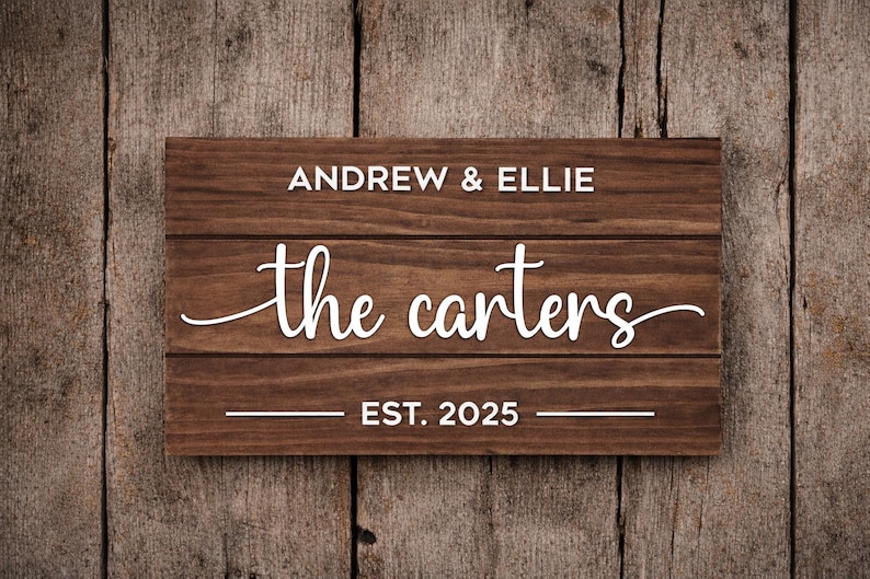 May include: A rectangular wooden sign with the names "Andrew & Ellie" above "the Carters" in a cursive font, and "EST. 2025" below. The sign is made of dark brown wood with visible grain, set against a wooden background.