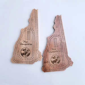 May include: Two wooden cribbage boards shaped like the state of New Hampshire. The boards are light and dark brown, with drilled holes for pegs and the text "New Hampshire: The Granite State" and a moose graphic.