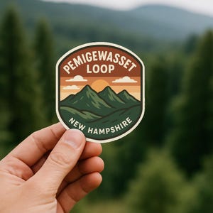 May include: A sticker with a landscape design featuring the text "PEMIGEWASSET LOOP NEW HAMPSHIRE". The sticker has a brown border, with a green and orange mountain scene, and a yellow sun.