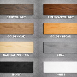 May include: A collection of eight rectangular wood samples showcasing various finishes. The samples include Dark Walnut, American Walnut, Golden Oak, Golden Pecan, Natural (No Stain), Gray, Ebony, and White, each labeled with its name.