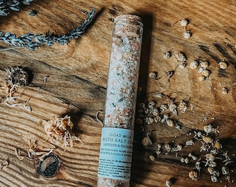 May include: A clear glass tube filled with bath salts, lavender buds, and other botanicals, with a cork stopper. The tube is labeled "Goat Milk Bath Salt Soaks Lavender & Buckwheat". The tube is on a wooden surface with dried flowers and lavender sprigs.