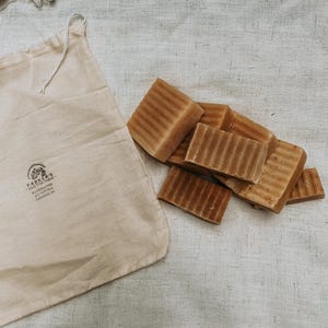 May include: A pile of brown handmade soap bars in a white cotton muslin bag. The bag has a black stamp that reads "Parker's Old Mill Farm Handmade All Natural  Cazenovia, NY".