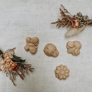 Puede incluir: Cuatro barras de jabón hechas a mano con forma de mariposa, mariquita, flor y oso. Los jabones son de color beige claro y están dispuestos sobre un fondo de tela blanca.
