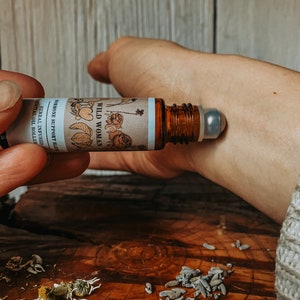 Könnte beinhalten: Eine braune Glas-Rollflasche mit einem silbernen Rollball. Das Etikett ist weiß mit einem floralen Design und dem Text "WILD WOMAN HORMONES SUPPORT BLEND HERBAL OIL INFUSED ESSENTIAL OIL ROLLER".