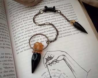 Könnte beinhalten: Eine goldene Kette mit einem schwarzen und orangefarbenen Kristallpendel hängt über einem Buch mit Text über Intuition und das Unterbewusstsein. Das Pendel wird für Radiästhesie und Wahrsagerei verwendet.