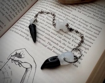 Könnte beinhalten: Zwei Pendel mit schwarzen und weißen Kristallspitzen, die an einer silbernen Kette hängen. Die Pendel liegen auf einem Buch mit Text über Rutengehen und Intuition.