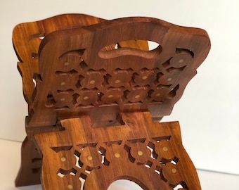 Op de afbeelding: Een houten boekenstandaard met ingewikkelde houtsnijwerk en messing accenten. De standaard is gevouwen en heeft een bruine afwerking.