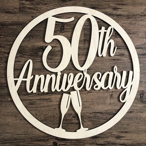 May include: A white wooden circle with the text "50th Anniversary" and two champagne glasses.