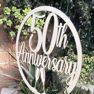 May include: A round wooden sign with the text "50th Anniversary" in a decorative font. Two champagne glasses are depicted below the text.