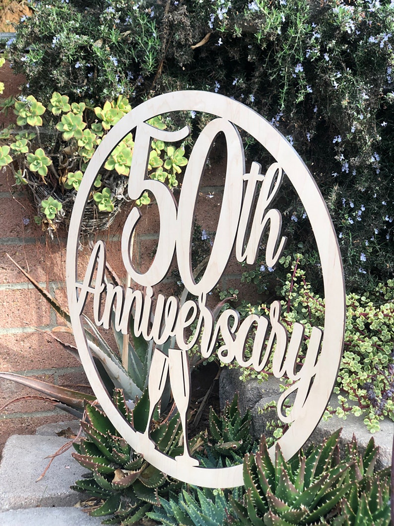 May include: A round wooden sign with the text "50th Anniversary" in a decorative font. Two champagne glasses are depicted below the text.