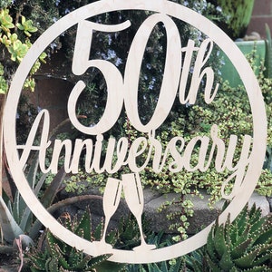 May include: A wooden circle sign with the text "50th Anniversary" and two champagne glasses cut out. The sign is light brown and has a natural wood grain.