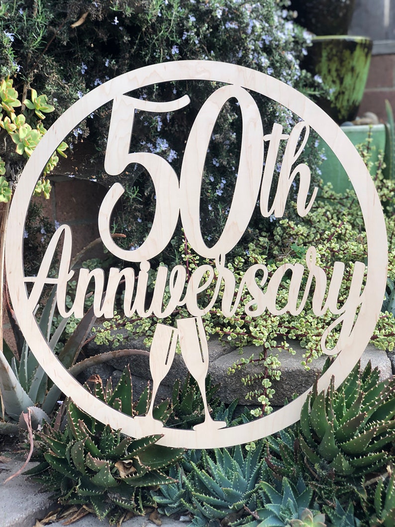 May include: A wooden circle sign with the text "50th Anniversary" and two champagne glasses cut out. The sign is light brown and has a natural wood grain.