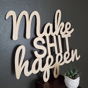 May include: Wooden sign with the text "Make Shit Happen" in cursive lettering. The sign is mounted on a dark gray wall and a small potted succulent plant is in the foreground.