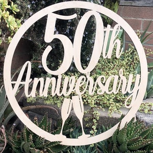May include: A wooden circle sign with the text "50th Anniversary" and two champagne glasses cut out. The sign is light brown and has a rustic look.