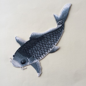 Può includere: Una toppa da stirare ricamata grigia a forma di pesce koi con accenti bianchi. Il pesce nuota verso destra con la pinna caudale sollevata.