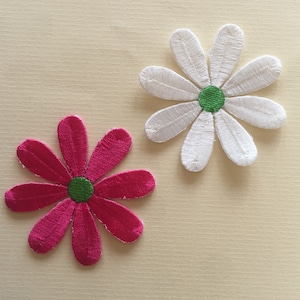 Può includere: Due fiori di stoffa ricamati, uno bianco e uno rosa, con centri verdi. I fiori sono su uno sfondo beige chiaro.