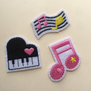 以下が含まれることがあります： 音楽をテーマにした刺繍のアイロン接着パッチが3つ。1つのパッチは、ピンクのハートが付いた白黒のピアノ、もう1つは、黄色の星が付いたピンクの音楽記号、最後は、ピンクの音楽記号と黄色のG記号が付いた白黒の五線譜です。