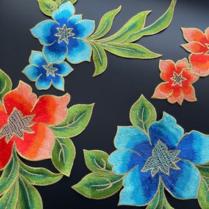 May include: Embroidered floral appliques in shades of blue and orange with green leaves and gold outlines. The appliques feature detailed stitching and are arranged on a dark surface. These decorative patches are ideal for fabric embellishment.