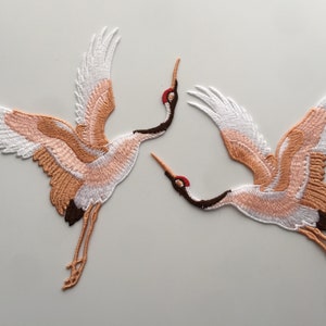 May include: Two embroidered iron-on patches of white and peach cranes with red beaks and brown legs. The cranes are in flight with their wings spread.