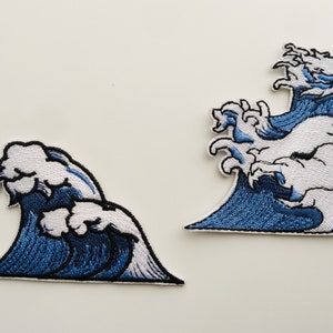 Puede incluir: Dos parches bordados para planchar que representan olas azules y blancas. El parche más grande presenta una ola grande con una cresta que se curva.