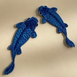 Könnte beinhalten: Zwei blaue gestickte Fisch-Patches mit einem schuppigen Design. Die Fische sind in entgegengesetzte Richtungen gerichtet und haben einen leicht dreidimensionalen Effekt.