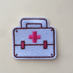 May include: White embroidered patch of a first aid kit with a brown outline and a pink plus sign in the centre.