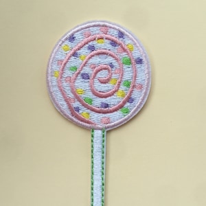 May include: An iron-on patch embroidered with a white and pink lollipop, featuring a green stick. The lollipop has a pink swirl and is decorated with purple, yellow, and green dots.