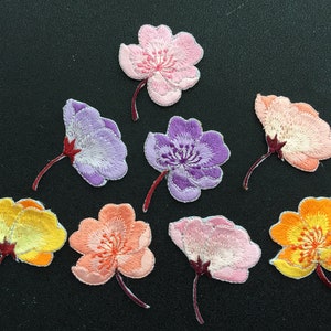 May include: Assortment of eight embroidered flower patches in various colors, including pink, purple, orange, and yellow. The patches are all in the shape of a flower with five petals.
