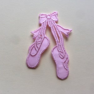 May include: Pink fabric ballerina shoe applique with a bow. The shoes are tied together with a ribbon.