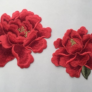 Puede incluir: Dos parches de flores de tela bordados en rojo. El parche más grande es una sola flor, mientras que el parche más pequeño tiene una sola flor con dos hojas verdes.