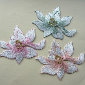 Op de afbeelding: Drie geborduurde bloemvlekken in roze en groene tinten. De bloemen hebben witte bloemblaadjes met roze of groene randen en gouden centra. De vlekken liggen op een lichtbeige achtergrond.