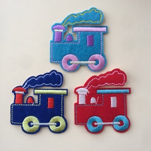 Può includere: Tre toppe per treni in feltro colorate. Una è blu con accenti viola e rosa, un'altra è blu navy con accenti gialli e rossi, e l'ultima è rossa con accenti blu e bianchi. Ogni treno ha dettagli ricamati e una nuvola di fumo.