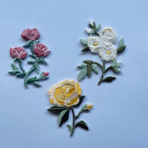 Könnte beinhalten: Drei gestickte Bügelbilder mit Blumenmotiven. Das erste Bügelbild ist rosa und zeigt drei Rosen. Das zweite Bügelbild ist weiß und zeigt drei Blumen. Das dritte Bügelbild ist gelb und zeigt eine einzelne Rose.