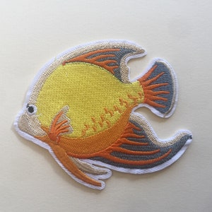 May include: Embroidered patch depicting a yellow fish with orange and grey details. The fish has a white border and a detailed design, suitable for application on clothing or accessories. The patch is a decorative item.