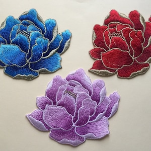 Puede incluir: Tres parches de flores de tela bordados en azul, rojo y morado. Las flores tienen todas el mismo diseño, pero de diferentes colores.