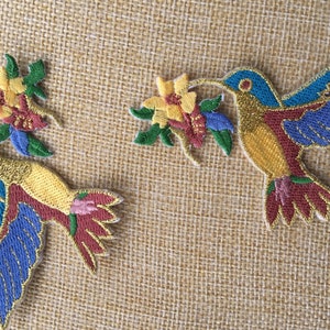 May include: Two embroidered iron-on patches featuring hummingbirds with outstretched wings, each holding a yellow flower in their beaks. The hummingbirds are a mix of gold, blue, red, and green.