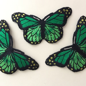 Op de afbeelding: Drie groene geborduurde vlinderpatches met gele accenten. De vlinders zijn in een driehoekige vorm gerangschikt.