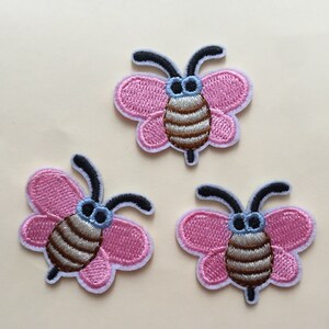 Puede incluir: Tres parches bordados de abejas rosas y marrones con antenas negras y ojos azules.