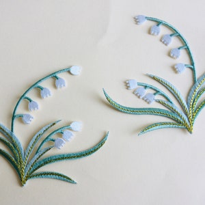 Peut inclure: Deux patchs brodés de fleurs de muguet blanches avec des feuilles vertes et des accents dorés.