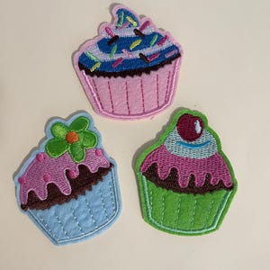 May include: Three embroidered cupcake patches. One is pink with blue and white frosting and sprinkles. One is light blue with pink frosting and a green flower. One is green with pink frosting and a red cherry.