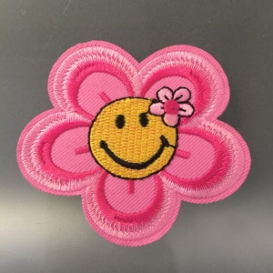 Könnte beinhalten: Rosa blumige Patch mit einem gelben Smiley-Gesicht und einer rosa Blume oben drauf.