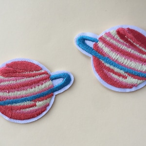 May include: Two embroidered planet patches. Each patch features a red and pink striped planet with a blue ring, set against a white background. The patches are designed with a whimsical, cartoonish aesthetic, ideal for adding to clothing or accessories.